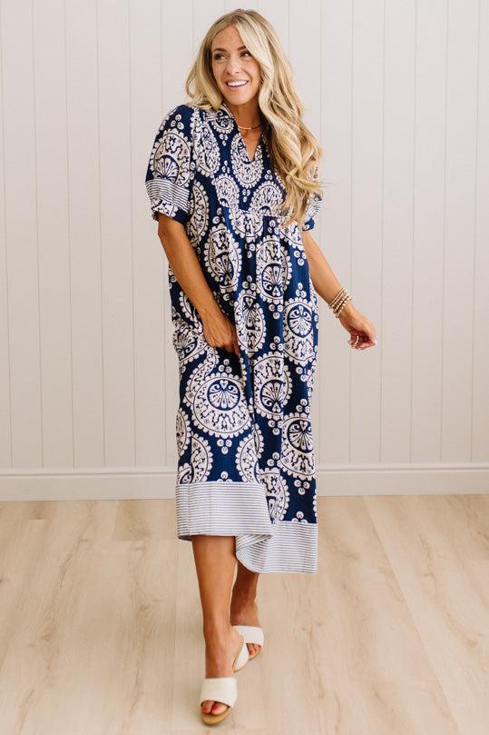 A woman modeling a navy blue and white paisley midi dress with a striped hem, standing in a room with light wood floors and white paneled walls.