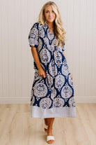 A woman modeling a navy blue and white patterned midi dress with short sleeves, a V-neckline, and a striped hem, standing in front of a white paneled wall and light wood floor.