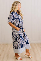 A woman modeling a navy blue and white paisley midi dress with striped trim, standing in a room with light wood floors and white paneled walls.