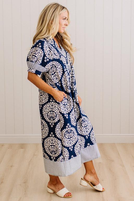 A woman modeling a navy blue and white paisley midi dress with striped trim, standing in a room with light wood floors and white paneled walls.