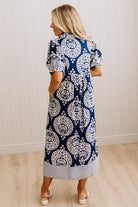 A woman wearing a navy blue and white patterned midi dress, shown from the back in a room with white paneled walls and light wood floors.