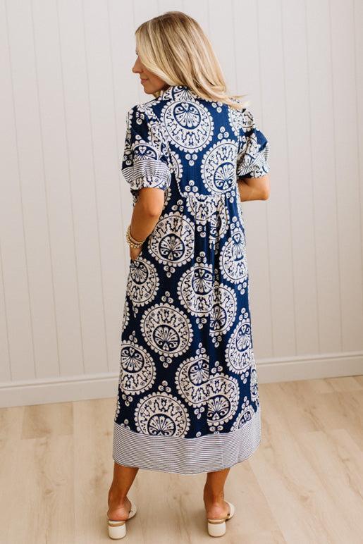 A woman wearing a navy blue and white patterned midi dress, shown from the back in a room with white paneled walls and light wood floors.