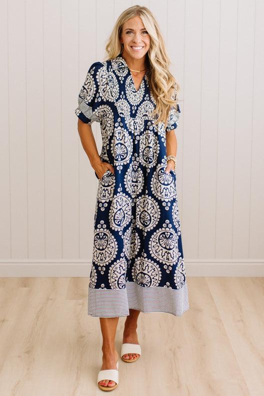 A woman modeling a navy blue and white paisley print midi dress with a striped hem, standing in a room with white paneled walls and light wood floors.