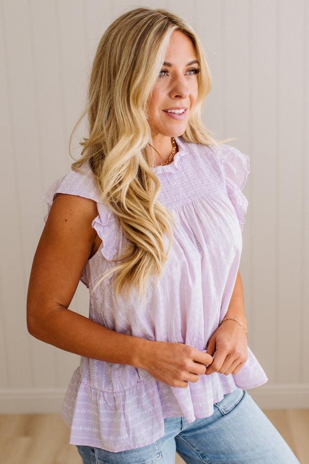Woman wearing a light purple top with ruffled sleeves and denim jeans against a neutral background