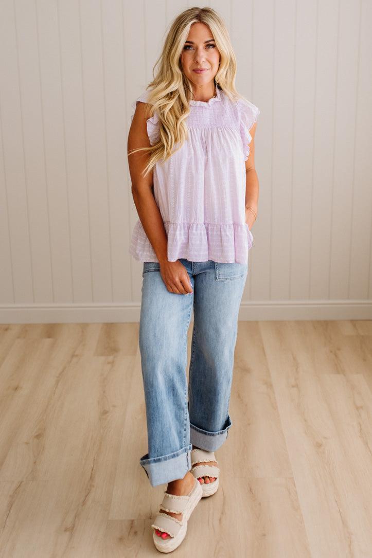 Woman wearing a light purple top and blue jeans standing on a wooden floor.