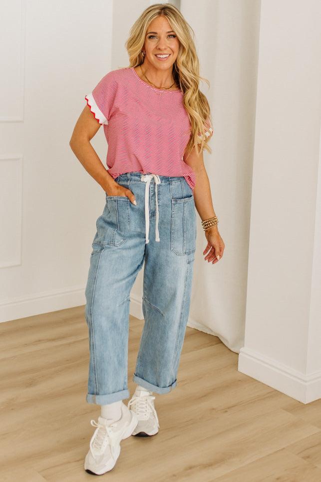 Woman wearing a pink top and blue jeans standing in a room with wooden flooring and white walls.