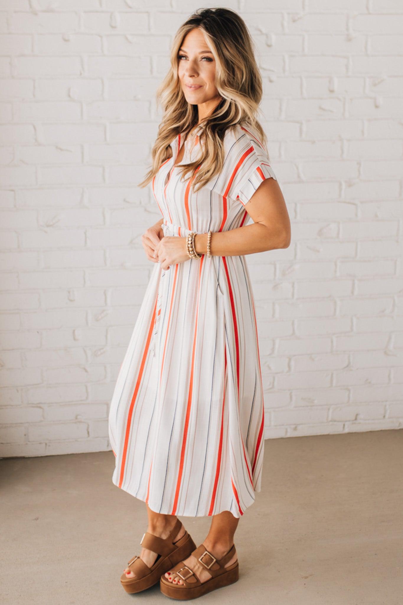 BLONDE WOMAN WEARING A RED AND WHITE DRESS WITH VERTICAL STRIPES, SIDE POCKETS, SHORT SLEEVE, BUTTON DOWN, FAUX TIE WAIST AND MIDI LENGTH.