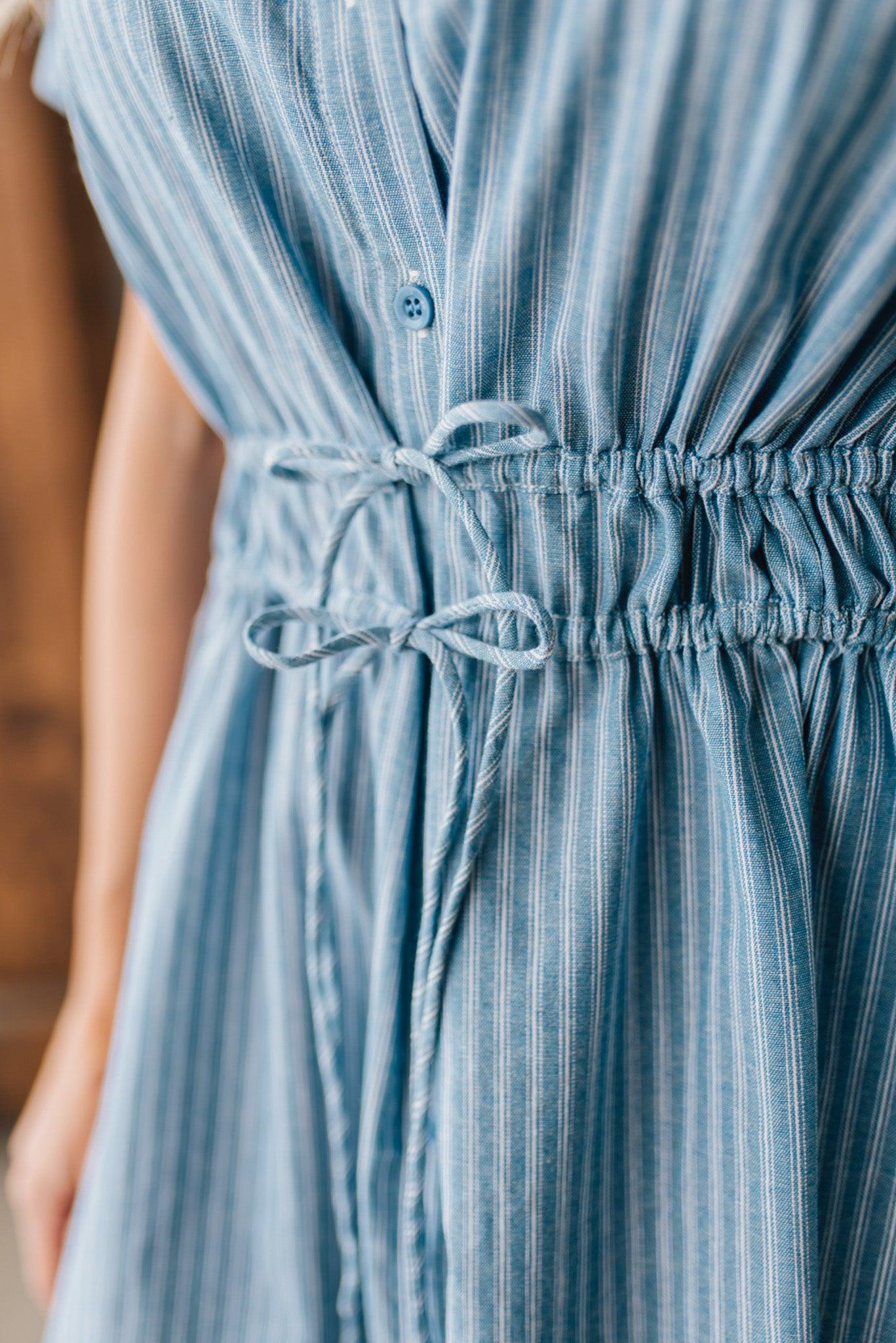 Blonde woman wearing a blue mid lenght dress with button front, double drawstring waist, and collar.