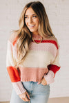 Blonde woman wearing a chunky waist length sweater with pink, mocha, red and cream colors.