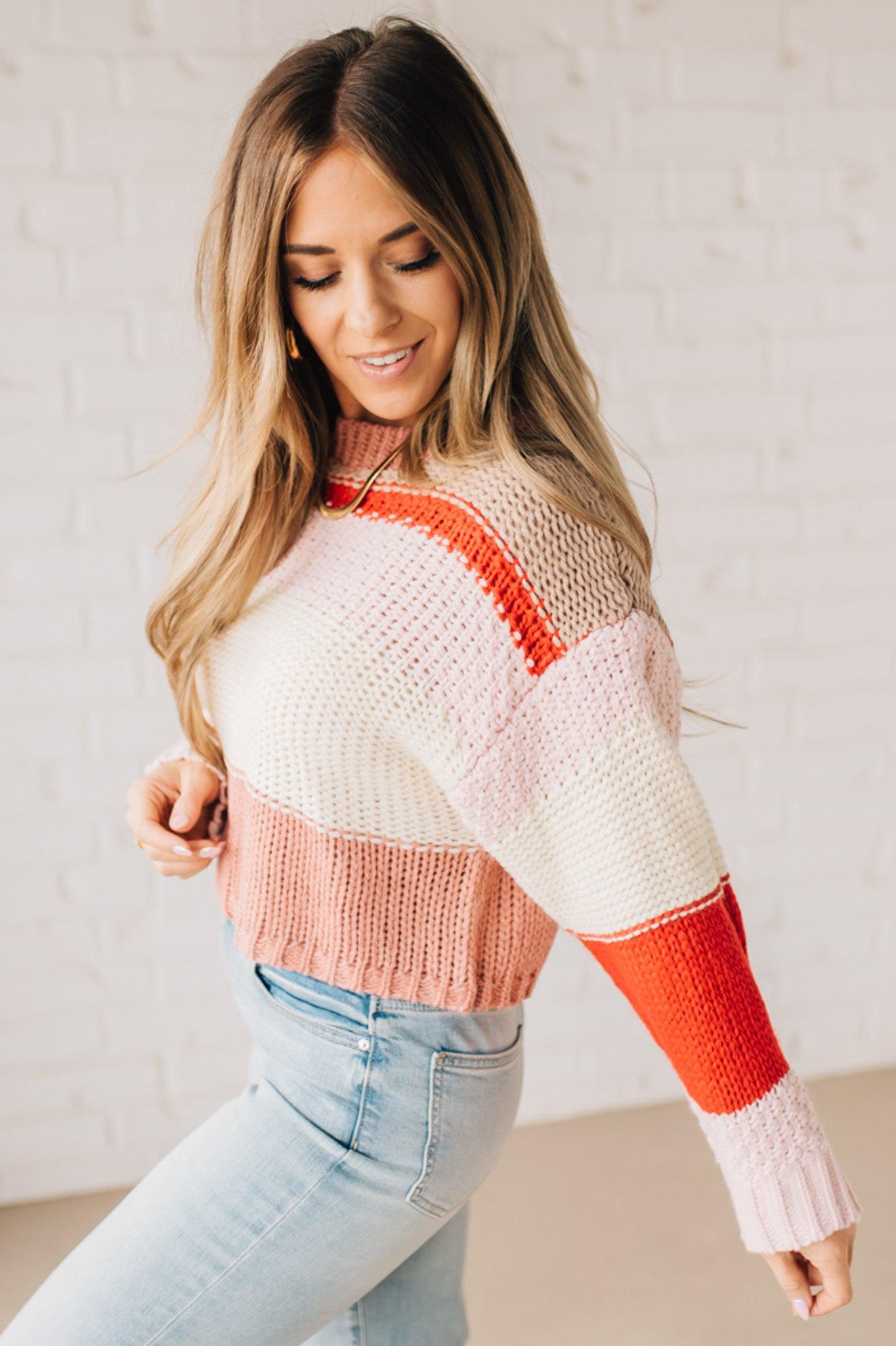 Blonde woman wearing a chunky waist length sweater with pink, mocha, red and cream colors.