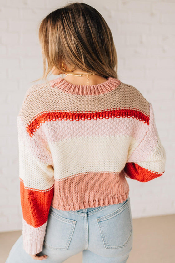 Blonde woman wearing a chunky waist length sweater with pink, mocha, red and cream colors.