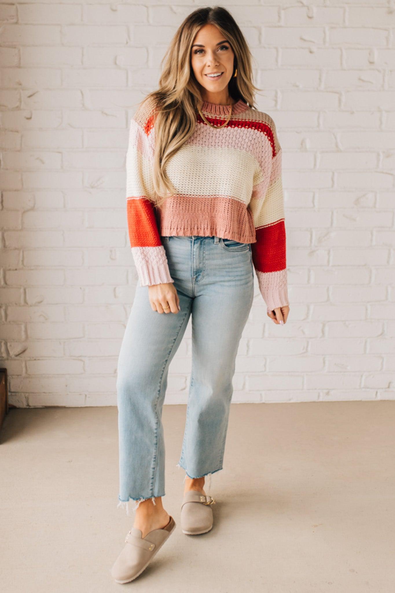 Blonde woman wearing a chunky waist length sweater with pink, mocha, red and cream colors.