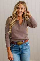 A smiling blonde woman wearing a brown and beige horizontally striped long-sleeve henley shirt, blue jeans, and a brown belt with a large gold buckle, standing against a white paneled wall.