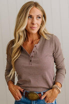A woman with blonde hair wearing a brown and beige striped long-sleeve henley shirt, blue jeans, and a large belt buckle, posing against a white paneled wall.