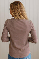 A woman viewed from behind wearing a long-sleeve top with thin horizontal brown and white stripes, paired with blue jeans, set against a light beige paneled wall.
