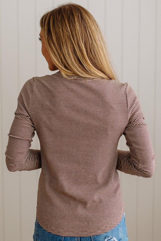 A woman viewed from behind wearing a long-sleeve top with thin horizontal brown and white stripes, paired with blue jeans, set against a light beige paneled wall.