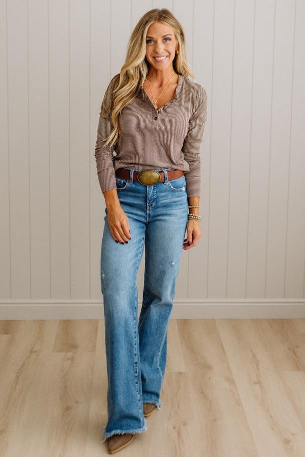 A woman modeling a brown long-sleeve henley top and light blue flare jeans with a brown leather belt, standing in front of a white paneled wall and light wood floor.