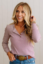 A smiling blonde woman wearing a pink and white striped long-sleeve henley top, blue jeans, and a brown belt with a large oval buckle, posed against a light gray paneled wall.