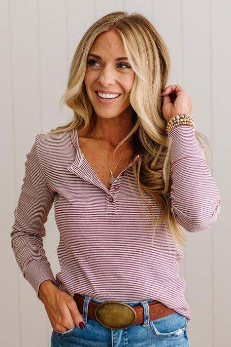 A smiling blonde woman wearing a pink and white striped long-sleeve henley top, blue jeans, and a brown belt with a large oval buckle, posed against a light gray paneled wall.