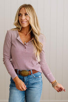 A woman modeling a long-sleeve henley top with thin pink and white horizontal stripes, paired with blue jeans and a brown belt, standing against a light beige paneled wall.
