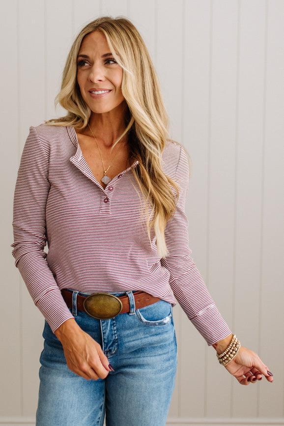 A woman modeling a long-sleeve henley top with thin pink and white horizontal stripes, paired with blue jeans and a brown belt, standing against a light beige paneled wall.
