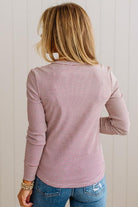 A woman viewed from behind wearing a dusty rose pink and white horizontally striped long-sleeve top with blue jeans, standing against a white paneled wall.