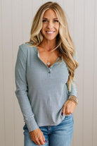 A smiling blonde woman wearing a light blue and white horizontally striped long-sleeve henley top with blue jeans, standing against a light beige paneled wall.