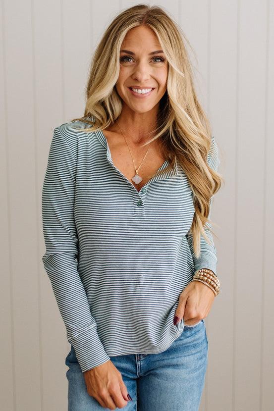 A smiling blonde woman wearing a light blue and white horizontally striped long-sleeve henley top with blue jeans, standing against a light beige paneled wall.