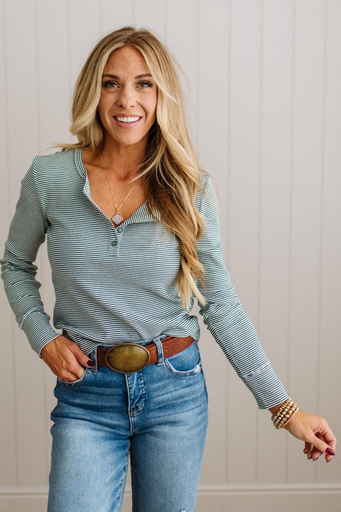 A smiling blonde woman wearing a light blue and white striped long-sleeve henley shirt tucked into blue jeans with a brown leather belt featuring a large oval buckle, standing against a white paneled wall.