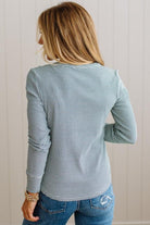 A woman shown from the back wearing a long-sleeve striped top in a muted blue-gray color with thin white stripes, paired with blue jeans, against a light beige paneled wall.