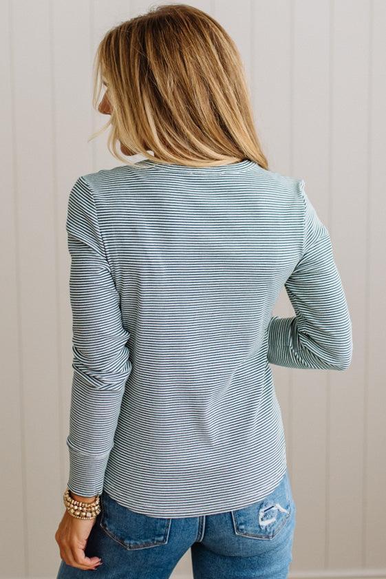 A woman shown from the back wearing a long-sleeve striped top in a muted blue-gray color with thin white stripes, paired with blue jeans, against a light beige paneled wall.