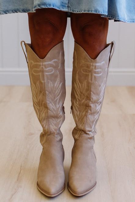 Beige cowboy boots worn with a denim skirt on a light wooden floor.