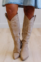 Beige knee-high boots worn with a denim skirt on a light wooden floor.