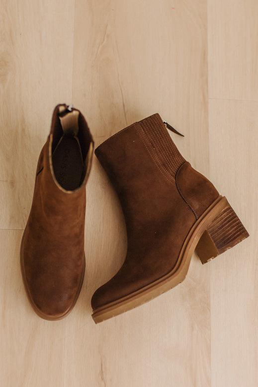 A pair of brown suede ankle boots with low stacked block heels, shown from a top-down view on a light wood plank floor.