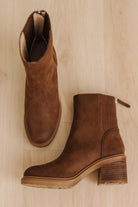 A pair of brown suede ankle boots with elastic side panels and stacked block heels, displayed on a light wood floor.