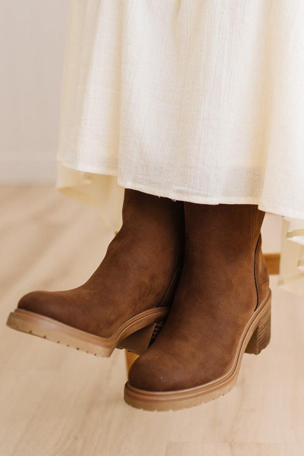 A close-up of a person wearing brown suede ankle boots with chunky block heels, paired with a flowing cream-colored skirt, standing on a light wooden floor.