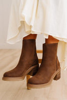 A pair of dark brown suede ankle boots with chunky block heels, worn with a flowing white dress on a light wood floor.