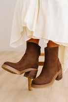A pair of women's brown suede ankle boots with chunky block heels, shown on a model's feet with a cream-colored skirt.