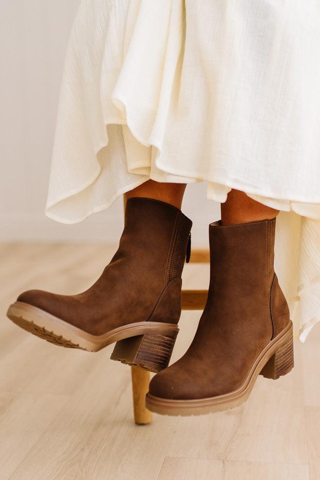 A pair of women's brown suede ankle boots with chunky block heels, shown on a model's feet with a cream-colored skirt.