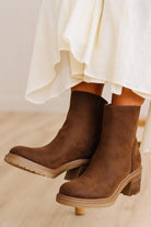 A pair of brown suede ankle boots with chunky block heels, shown on a model wearing a white skirt against a light wood floor background.