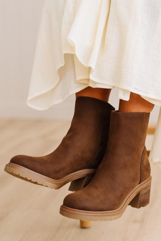 A pair of brown suede ankle boots with chunky block heels, shown on a model wearing a white skirt against a light wood floor background.