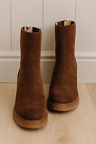 A pair of brown suede Chelsea-style ankle boots with elastic side gore panels and tan soles, standing on a light wood floor against a white paneled wall.