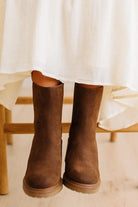 A close-up of a person's legs wearing brown suede ankle boots, with a white dress hem and a wooden chair visible in the background.