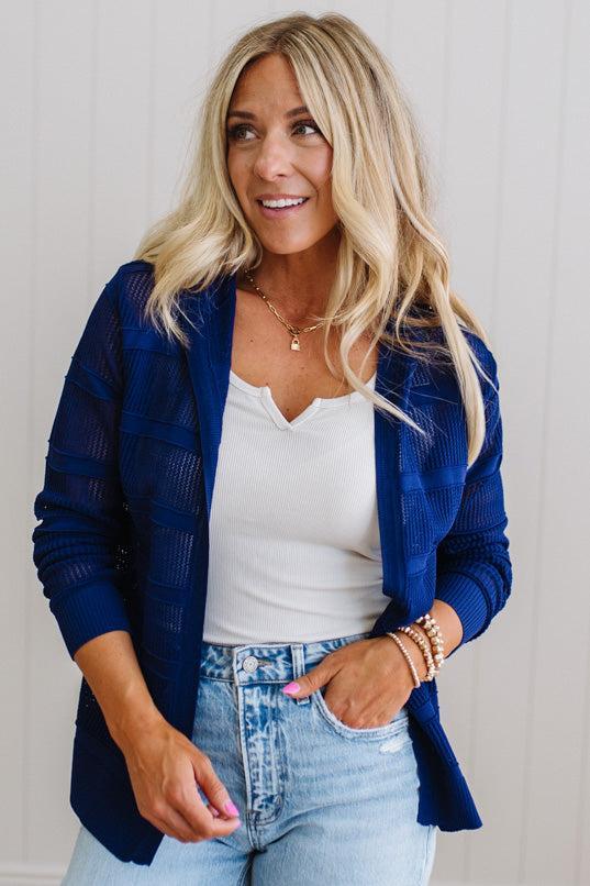 Woman wearing a blue cardigan, white top, and light blue jeans against a neutral background