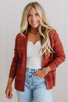 Woman wearing a rust-colored cardigan over a white top and blue jeans, standing against a light background.