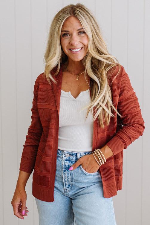 Woman wearing a rust-colored cardigan over a white top and blue jeans, standing against a light background.