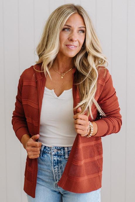 Woman wearing a rust-colored cardigan over a white top and blue jeans, standing against a light background.