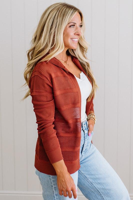 Woman wearing a rust-colored cardigan over a white top and light blue jeans against a neutral background