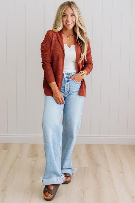 Woman wearing a rust-colored cardigan, white top, and light blue jeans against a white wall.