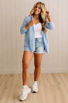 Woman wearing a light blue plaid shirt, white top, denim shorts, and white sneakers in a room with wooden flooring and a beige wall.